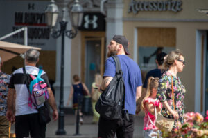 Фото тату на руке мужчины, идущего по улице. Татуировка выполнена в темных тонах, изображает детализированный рисунок. Мужчина одет в кепку и синюю футболку.