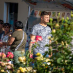 Фото тату на руке мужчины, изображающее лицо монахини в черном головном уборе, на фоне яркого киоска с мороженым и цветущих кустов.