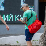 Фото мужчины идущего по улице с татуировкой на руке и красной спортивной сумкой через плечо. На заднем плане виден рекламный щит.