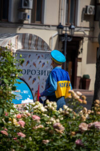 Фотография мужчины в форме с желтой повязкой "Волонтер" и флагом Украины, идущего по улице. На фоне виден киоск с мороженым.