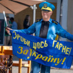 Фотография мужчины в форме с флагом и мечом, идущего по городской улице. Яркий солнечный день, городская архитектура на фоне.