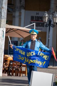 Фотография мужчины в форме с флагом и мечом, идущего по городской улице. Яркий солнечный день, городская архитектура на фоне.