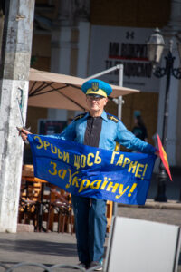 Фотография мужчины в голубой форме с украинской символикой, держащего сине-желтый флаг и саблю, идущего по улице.