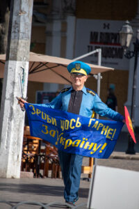 Фотография мужчины в голубой форме с украинской символикой, держащего сине-желтый флаг и саблю, идущего по улице.