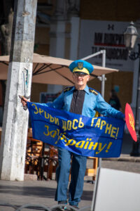 Фотография мужчины в голубой форме с украинской символикой, держащего сине-желтый флаг и саблю, идущего по городской улице.