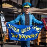 Фотография мужчины в голубой форме с украинской символикой, держащего сине-желтый флаг и саблю, идущего по городской улице.