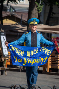 Фотография мужчины в голубой форме с украинской символикой, держащего сине-желтый флаг и саблю, идущего по городской улице.