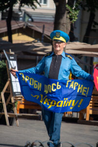 Фотография мужчины в голубой форме и фуражке с украинской символикой, держащего сине-желтый флаг и саблю на улице.