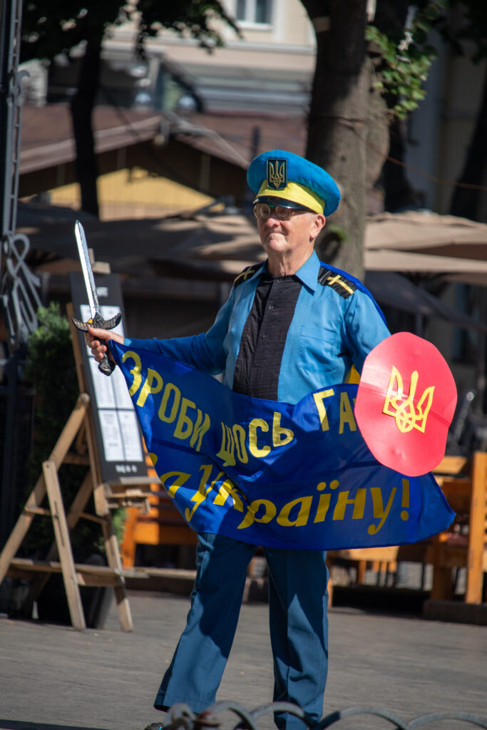 пожилой мужчина в одежде Супергероя стилизованное под флаг Украины и надписью сделай что-нибудь полезное для Украины5050