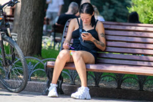 Фотография девушки, сидящей на скамейке и смотрящей в телефон. На плече и руке видны татуировки с растительным мотивом.