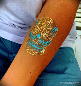 Фото тату: золотой череп в стиле сахарной черепушки (calavera) на руке. Детализированный рисунок татуировки с использованием ярких цветов и узоров.