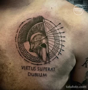 Фото татуировки шлема спартанца в круге с латинской надписью 'Virtus Superat Dubium' на груди. Татуировка выполнена в черно-серых тонах с использованием теней для создания объема.