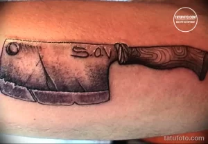 Фото черно-серой татуировки с изображением большого тесака и надписью 'S.oV'. Реалистичный рисунок на руке.