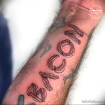 Фото татуировки на предплечье с надписью 'BACON', буквы которой выполнены из реалистичных изображений полосок бекона.