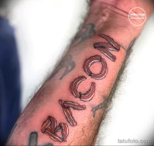 Фото татуировки на предплечье с надписью 'BACON', буквы которой выполнены из реалистичных изображений полосок бекона.