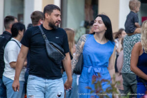 Фото пары: мужчина в черной футболке и джинсовых шортах, женщина в голубом платье с татуировкой на руке. Уличная фотография, демонстрирующая татуировки в повседневной жизни.