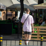 Фотография мужчины с татуировками на руках, стоящего спиной к камере на фоне летнего кафе. Современный городской стиль.