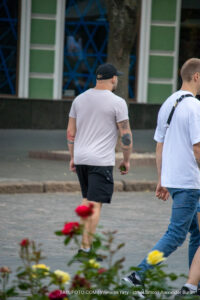 Фото тату: мужчина идет по улице, на его плече видна татуировка с изображением гор и леса. Черно-серый рисунок татуировки.