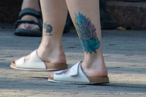 Фото тату: красочная татуировка в виде ловца снов на лодыжке девушки. Рисунок татуировки выполнен в акварельной технике с использованием ярких синих, голубых и розовых оттенков.