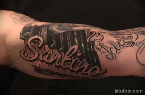 Фото тату на руке с изображением клавиш пианино и надписью "Santino". Детализированный рисунок татуировки в черно-серых тонах.
