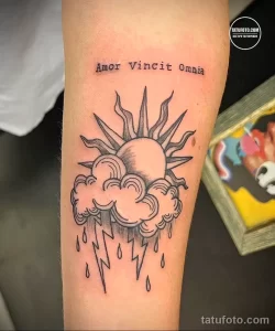 Фото тату на руке с изображением солнца, облаков и латинской фразы 'Amor Vincit Omnia', выполненное в технике тонких линий и минимализма.
