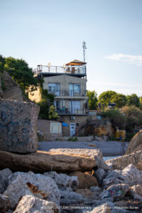 Фотография дома, расположенного на скале у моря. Закат, теплые тона, живописный пейзаж. На переднем плане камни и растительность.