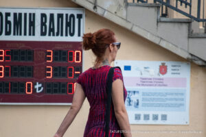 Фото тату на плече женщины на фоне табло с курсом валют. Рисунок татуировки выполнен в черных тонах, детализированный и привлекающий внимание.