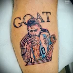 Фото тату с изображением Лионеля Месси, держащего кубок, и надписью "GOAT". Реалистичная проработка деталей, яркие цвета и динамичная композиция.