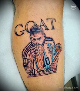 Фото тату с изображением Лионеля Месси, держащего кубок, и надписью "GOAT". Реалистичная проработка деталей, яркие цвета и динамичная композиция.