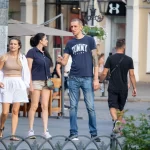 Фото мужчины с татуировкой на руке, одетого в синюю футболку и джинсы. Татуировка на плече выглядит как темный рисунок.