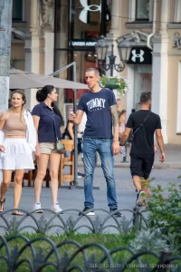 Фото мужчины с татуировкой на руке, одетого в синюю футболку и джинсы. Татуировка на плече выглядит как темный рисунок.