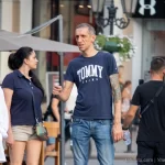 Фото мужчины с татуировкой на руке, одетого в синюю футболку и джинсы. Татуировка на плече выглядит как темный рисунок.