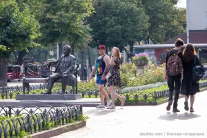 Фото тату на руке и ноге мужчины, идущего с девушкой в парке. Видны небольшие, но детализированные рисунки, выполненные в черном цвете.