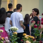 Фотография уличной татуировки на руке и ноге мужчины. Видны детализированные рисунки в стиле графика, выполненные черными чернилами. На заднем плане люди и цветы.