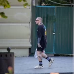Фотография мужчины с татуировками на руках и ногах, идущего по улице. Мужчина одет в черную футболку и шорты.