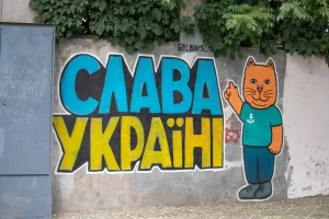 Фотография граффити на стене с надписью "Слава Украине!" и изображением оранжевого кота в зеленой футболке. Яркие цвета и простой стиль рисунка.