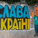 Фотография граффити на стене с надписью "Слава Украине!" и изображением оранжевого кота в зеленой футболке. Яркие цвета и простой стиль рисунка.