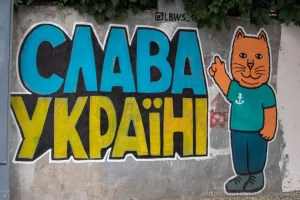 Фотография граффити на стене с надписью "Слава Украине!" и изображением оранжевого кота в зеленой футболке. Яркие цвета и простой стиль рисунка.