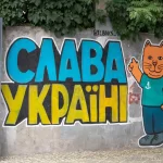 Фотография граффити на стене с надписью "Слава Украине!" и изображением оранжевого кота в зеленой футболке. Яркие цвета и простой стиль рисунка.