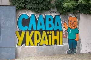 Фотография граффити на стене с надписью "Слава Украине!" и изображением оранжевого кота в зеленой футболке. Яркие цвета и простой стиль рисунка.