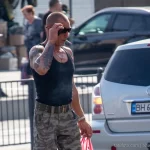 Фотография мужчины с татуировками на руке, курящего на улице. Вид сбоку, солнечный день, городской пейзаж.