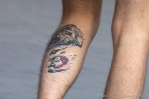 Фото тату на голени мужчины: изображение головы льва и человеческого лица, выполненное в черно-серых тонах. Видна часть обуви.