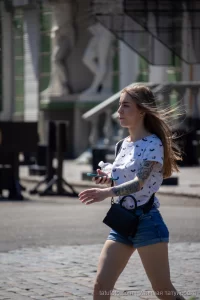 Фотография девушки в футболке и шортах с татуировкой на предплечье, идущей по улице в солнечный день.