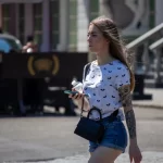 Фотография девушки в белой футболке и джинсовых шортах с татуировкой на предплечье, идущей по улице.