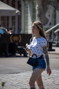 Фотография девушки в белой футболке и джинсовых шортах с татуировкой на предплечье, идущей по улице.