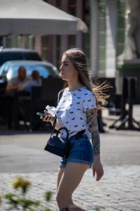 Фотография девушки в шортах и футболке с татуировкой на предплечье, идущей по улице в солнечный день.