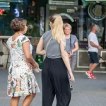 Фотография девушки с татуировкой на плече, изображающей цветы и бабочек в черно-серых тонах. Девушка идет по улице.