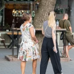 Фотография уличной татуировки на плече девушки, изображающей цветочный узор в черно-серых тонах. Девушка идет по улице с подругой.