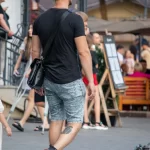 Фотография мужчины с татуировкой в виде спирали на икре ноги, идущего по улице с сумкой через плечо.