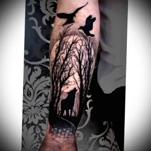 Фото татуировки в стиле blackwork на руке, изображающей волка, скрытого в лесу, над которым парит ворон, выполненной полностью в черном цвете.